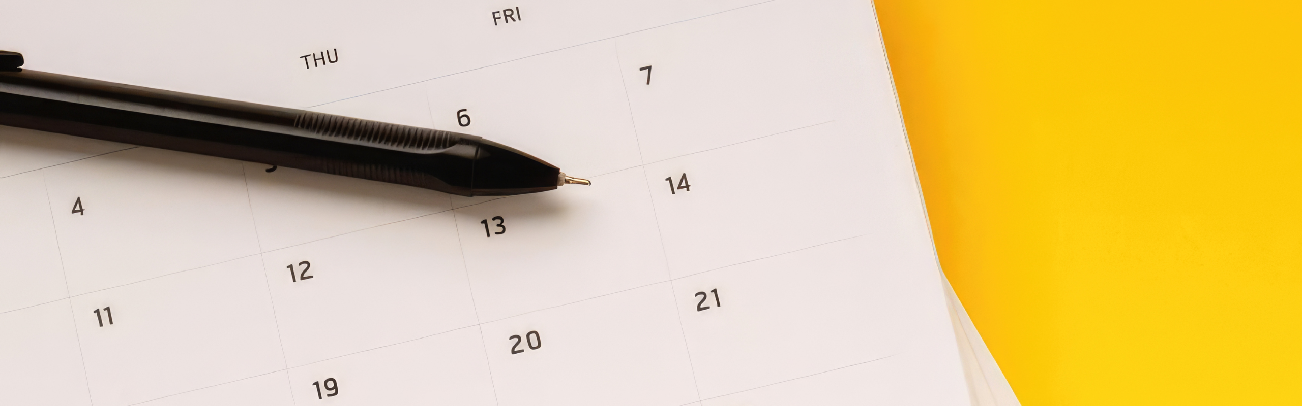 A black pen rests on an open calendar showing dates from Thursday the 6th through the 21st next to a yellow notebook.